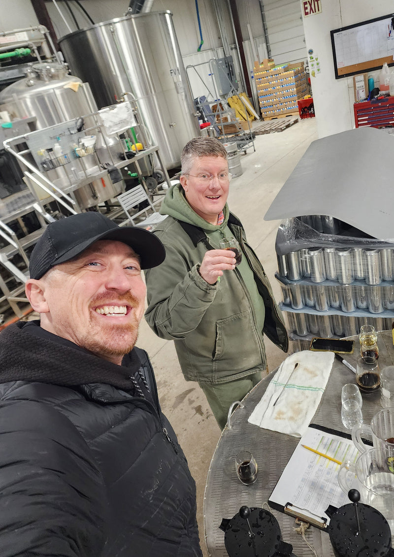 Two men smiling in a brewery workshop, with stainless steel tanks, brewing equipment, and stacks of cans visible in the background.