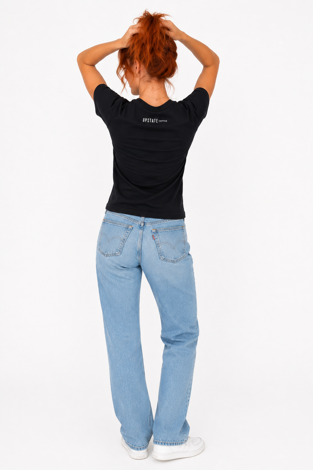 red hair Woman model showing off back of shirt holding hair up wearing a black 'Upstate Coffee'  t-shirt and light blue jeans on a white background