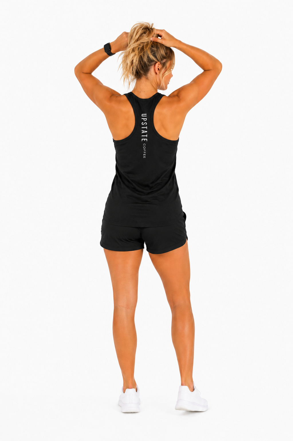 Woman model wearing a black tank top holding her hair up showing off the back of an upstate coffee mountain scene graphic design and black shorts on a white backdrop