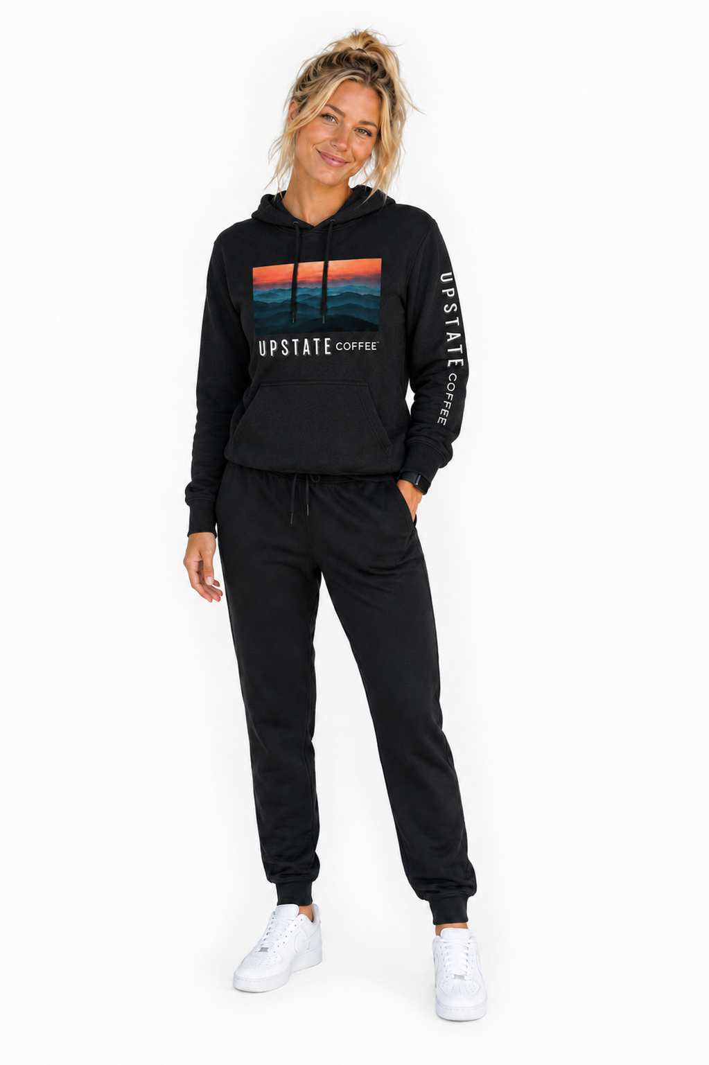 Blond model wearing a black hoodie and jogger set with 'Upstate Coffee' mountain scene branding on a white background